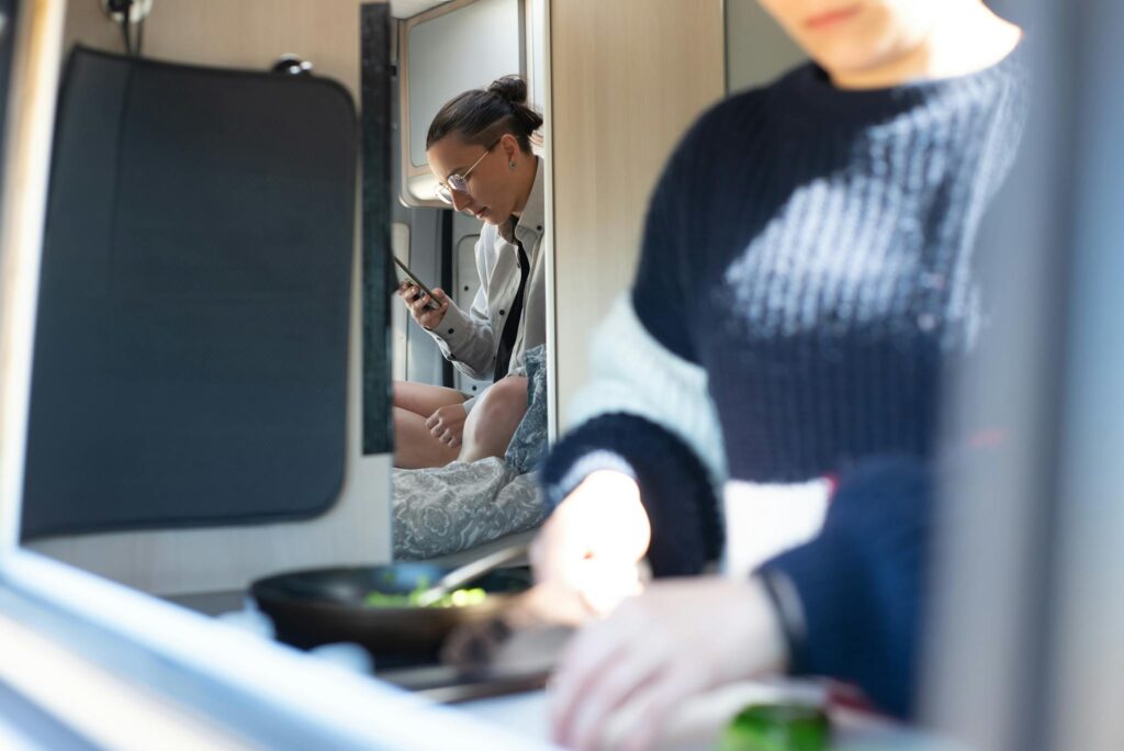 A woman multitasks in a cozy caravan, using a smartphone while cooking.