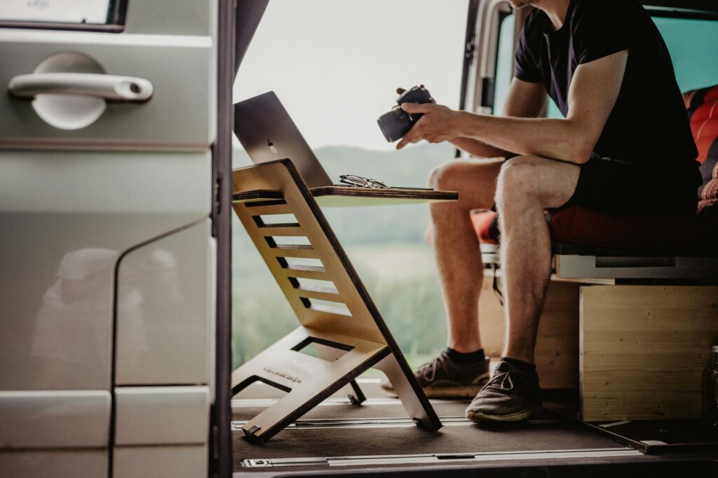 Man working remotely from a van with a portable desk, camera, and laptop setup, emphasizing mobile office lifestyle.